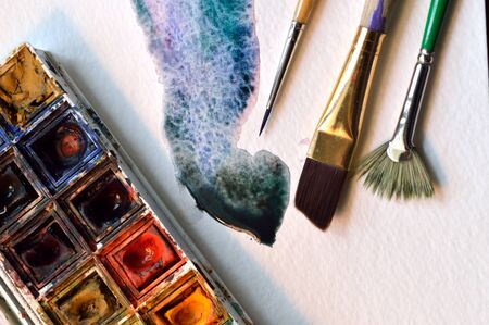 Used artist's watercolor palette, on watercolor paper with example of wet on wet technique of a fluid watercolor wash, with three brushesの写真素材