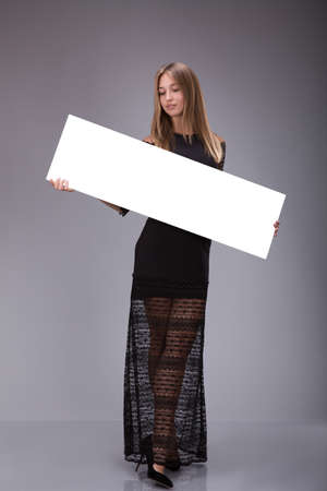 Young woman in black dress holding blank poster. Girl showing banner with copy space over gray backgroundの写真素材