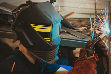 Male in face mask welds with argon-arc welding in body shop