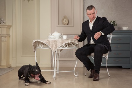 Man seatting at the table and looking at the watch. Near leas a dog pit bull or black Staffordshire Terrier in vintage interior. studio shootの写真素材