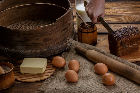 The hand cuts black bread on the background of ingredients for cooking bread. eggs, honey, milk,...