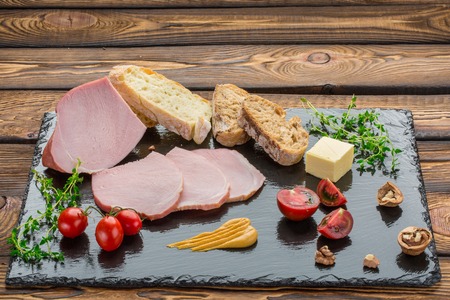 Pieces of cold-boiled, bread, butter, tomatoes, herbs, walnut, mustard. Ingredients for breakfast on black stone board over wooden backgroundの写真素材