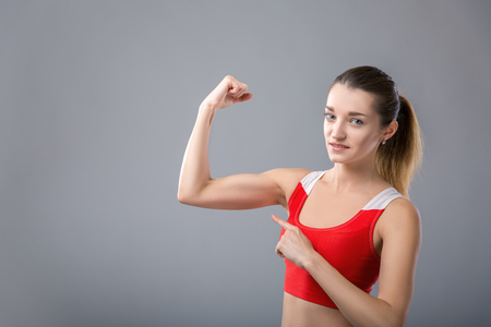 Beautiful stylish young woman is pointing on her muscles and smiling, on gray background. space for textの写真素材