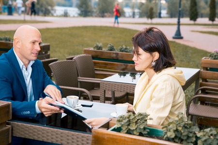 Woman communicates with bald man showing her something in the paper notebook at the desk at the summer cafe on the street of cityの写真素材