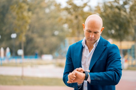 Serious male bald entrepreneur in formal suit looking at wristwatch planning working schedule , confident executive manager checking time while waiting for meeting standing outdoorsの写真素材