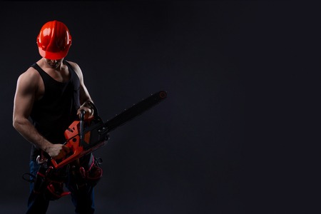Chainsaw and young worker in the helmet. The naked man broke the chainsaw. Attractive guy with tool on black background. Builder or lumberjack, wood processingの写真素材