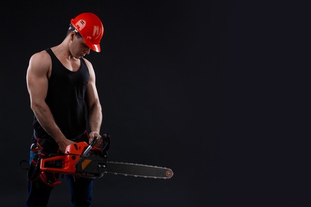 Chainsaw and young worker in the helmet. The naked man broke the chainsaw. Attractive guy with tool on black background. Builder or lumberjack, wood processingの写真素材