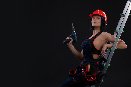Beautiful woman with a drill on a ladder ready to use it on black backgroundの写真素材