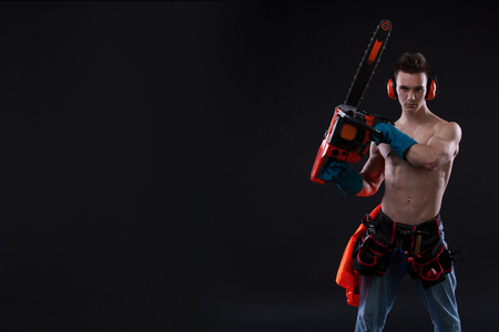 portrait of aggressive muscular man in headphones with chainsaw in hand, posing on black background. builder. Copy spaceの写真素材
