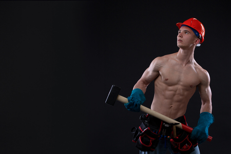 Handsome young topless construction worker in helmet with sledge hammer, studio shot, black background. builder. Copy spaceの写真素材