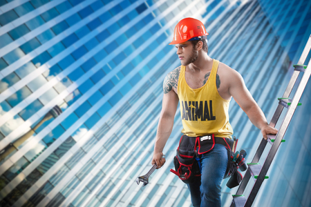 young handsome macho man builder with sexy muscular athletic strong body in orange hard hat or helmet holds wrench near wood ladder against the background of a glass skyscraper.の写真素材