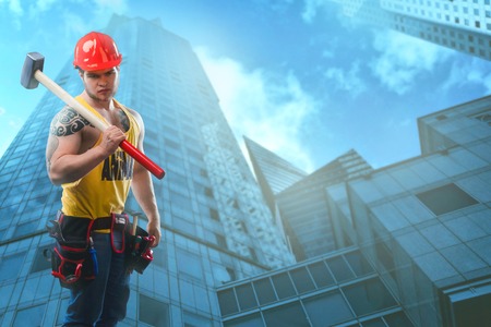 young handsome macho man builder with sexy muscular athletic strong body has bare torso holds hammer against the background of a skyscraper. Copy spaceの写真素材