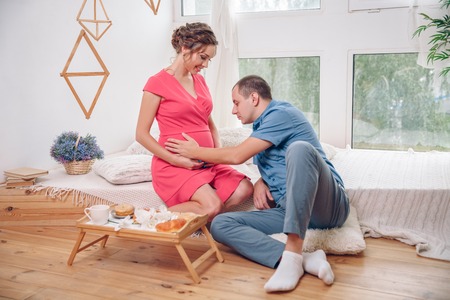 Happy pregnant woman with her husband are holding and listening to baby in belly and setting on living rooms floor at homeの写真素材
