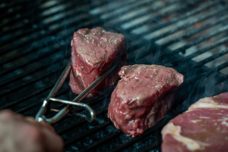Steak and rib are cooking on grill. Pork ribs and steak on hot grill. Restaurant kitchen, culinary. Tongs Holding Grilled Pork Ribsの写真素材