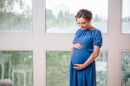 young pregnant woman staying near the window in blue dress. in anticipation of the childの写真素材