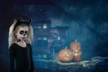 Halloween, holidays, masquerade concept - the portrait of young little beautiful girl with skull makeup and horns. Halloween, face-art, skull make up concept. on a background of pumpkins and an old house in the forestの写真素材