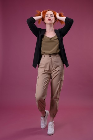 young redhead woman touching hair and looking at camera on pink backgroundの写真素材