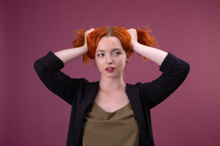 young redhead woman touching hair and looking at camera on pink backgroundの写真素材