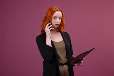 Portrait of a beautiful amazing redhead woman posing over pink background holding folder talking by mobile phone.の写真素材