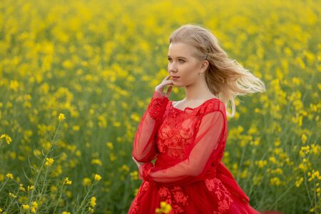 Portrait of blond teen girl wearing at red dress posing at field with yellow rapeseed flowers.の写真素材