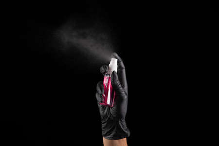 A female hand in a rubber black glove sprays a hand antiseptic from bottle on a black background....