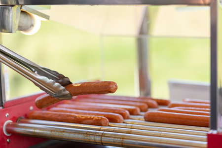 Greasy hot dog sausages on a culinary machine in a cafe. Cooking sausages for a hot dog bun with sesame seeds in the background. Traditional american fast food.の写真素材