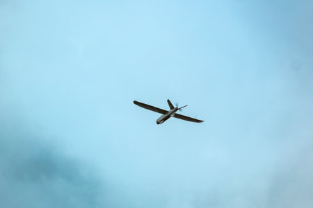 A drone flies against a bright blue sky with scattered clouds, surrounded by blurred green leaves in the foreground.の写真素材