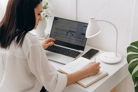 Back view of young freelancer woman sitting at white desk using laptop writing notes while studying online, watching webinar, looking at pc screen learning web classes or remote working from homeの写真素材