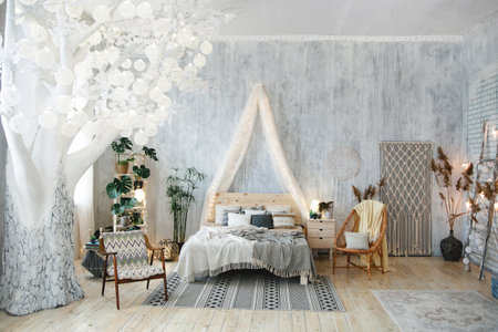 Elegant boho bedroom setup with white tree with cozy lights, soft bedding, woven chair, houseplants, light grey walls, and boho-chic accents. Room includes decorative rugs and artistic decor piecesの写真素材