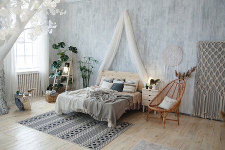Elegant boho bedroom with a white tree adorned with lights, soft bedding, wicker chair, houseplants, light grey walls, and boho-chic accents. Room features a decorative rug and artistic decor piecesの写真素材