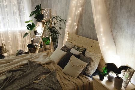 Boho bed with wooden headboard, soft blankets and pillows, surrounded by light grey walls, ladder shelf with plants, and fairy lights. The room has a cozy and warm atmosphereの写真素材