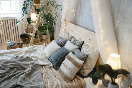Boho bed featuring wooden headboard, assorted pillows, soft blankets, light grey walls, houseplants, and fairy lights. Room exudes warmth and tranquility with a stylish touchの写真素材