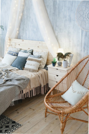 Stylish boho bed with wooden headboard, soft blankets, assorted pillows, and light grey walls. Accompanied by wicker chair, macrame wall hanging, bedside table, and decorative vase with dried plantsの写真素材