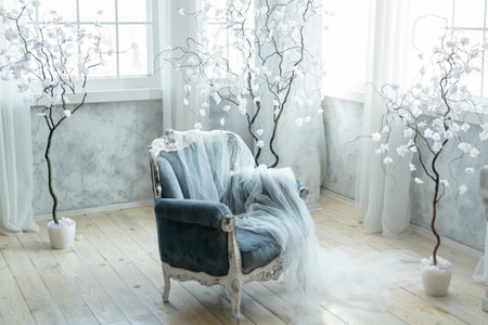 Elegant blue velvet armchair draped with sheer white fabric, surrounded by decorative white trees in a softly lit room. Creates luxurious and ethereal atmosphere, perfect for high-end interior designの写真素材