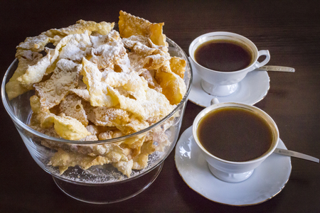 Sprinkle with powdered sugar, fried plate of shortcake dough. Dessert Polish and Lithuanian cuisine - faworki (angel wings). Still life. Faworki in a glass vase, coffee in a two porcelain cup with saucer and spoon on dark background.の写真素材