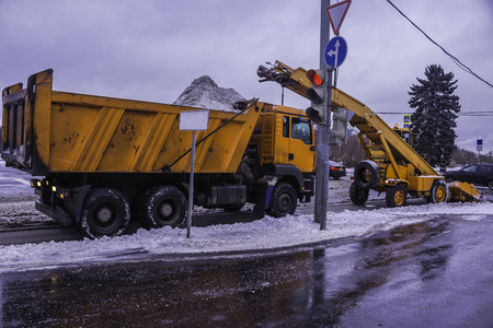 Machine shovels snow from the street and loading snow into a truck . Orange machines, snow cleaners , near traffic lights . Moscow , Russia .のeditorial素材
