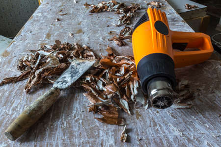Restoration of an old door. Removing old paint coats with a heat gun and a spatula. Gnarled pieces of brown paint and old white putty under the tools.の写真素材