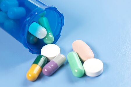 Colorful capsule and tablets spill from blue prescription container on blue background.