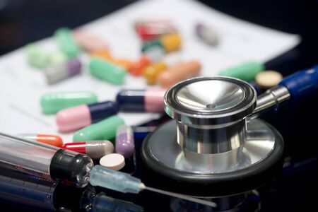 Stethoscope, syringe, different colored medications, and written doctor's prescription on dark reflective surface.の写真素材