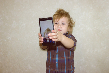 A boy kid looks into the camera of a smartphone, shows the screen with his digital photo. Toddler smiles at the camera and takes a selfie.の写真素材