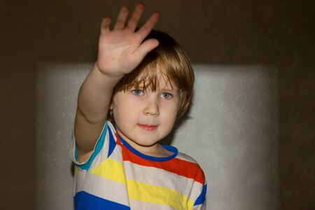 A little boy puts forward his hand showing a stop sign on the background of a rectangular frame of light from a film projector on the wall.の写真素材