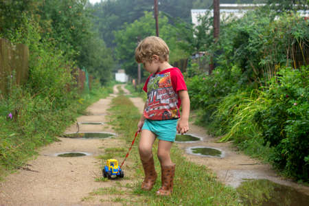 BEREZINO, BELARUS - AUGUST 13, 2019: little boy in rubber boots pulls a toy toy car tractor on a rope along a country sandy road with puddles after rain.のeditorial素材