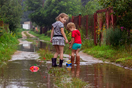 BEREZINO, BELARUS - AUGUST 13, 2019: two young children in rubber boots pull toys on a rope along a country sandy road with puddles after rain.のeditorial素材