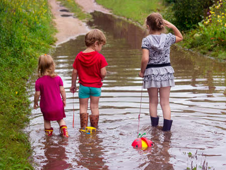 BEREZINO, BELARUS - AUGUST 13, 2019: three young children in rubber boots pull toys on a rope along a country sandy road with puddles after rain.のeditorial素材