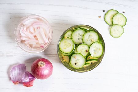 Sliced pickled cucumbers in a glass bowl with pickle onions on white wooden background
