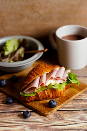 Fresh baked croissant sandwich with ham, cheese and lettuce on wooden board. Healthy breakfast.の写真素材