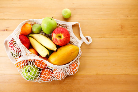 Fresh fruits in reusable grocery net bag on wooden table. Apple, banana, papaya, orange and lime in eco cotton bag. Zero waste, plastic-free and sustainable lifestyle.の写真素材