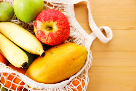 Fresh fruits in reusable grocery net bag on wooden table. Apple, banana, papaya, orange and lime in eco cotton bag. Zero waste, plastic-free and sustainable lifestyle.の写真素材