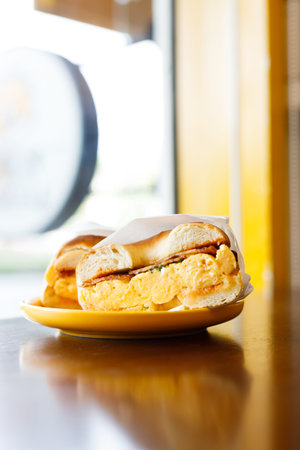 Bagel bread with scrambled egg and bacon slices on yellow dish. Bagel sandwich breakfast with egg and bacon in cafe.の写真素材