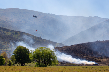 A helicopter dampens hot spots during a controlled scrub fire to clear farmland covered in gorse and bushの写真素材